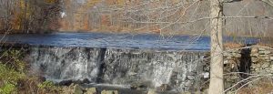 A wide, low-head stone dam with water cascading over a straight edge into a rocky creek bed.
